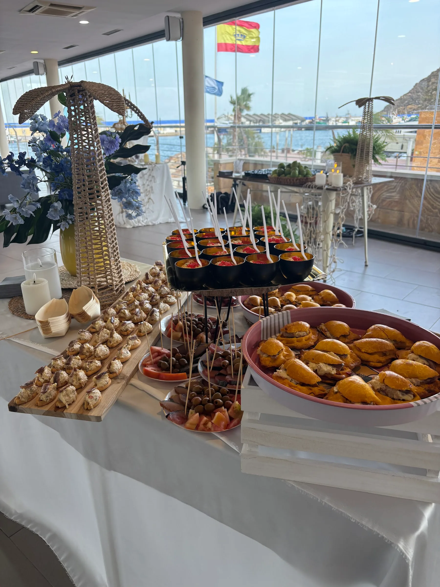 Mesa de evento montada en Mazmara Sunset con canapés y montaditos, y el puerto al otro lado del ventanal