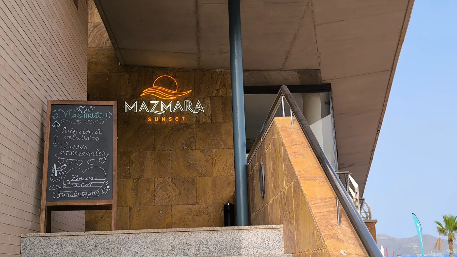 Entrada de Mazmara Sunset: el rótulo de neón con el sol sobre la ola y la pizarra con la carta del día